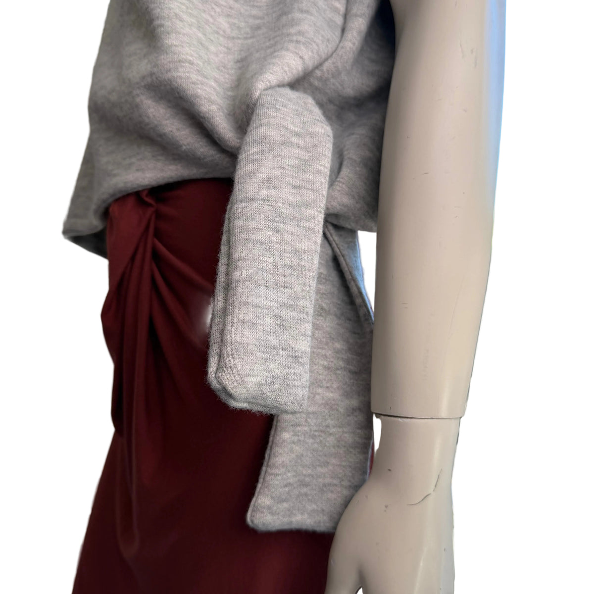 A close-up of a mannequin torso wearing a light gray knit top and a dark red skirt, with a window and outdoor view in the background.