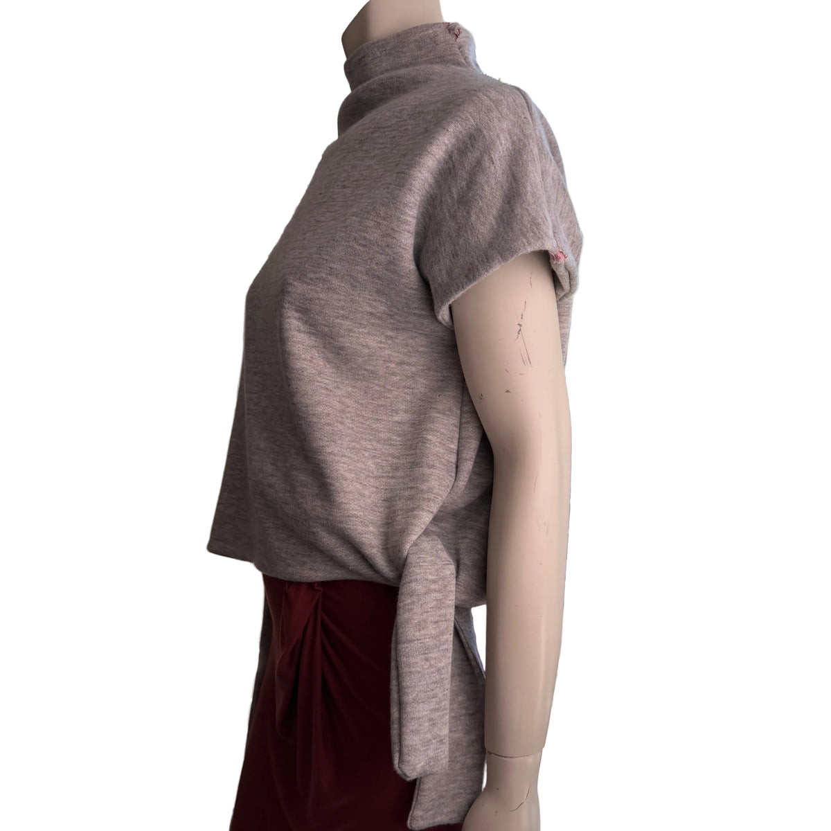 A taupe short-sleeve mock-neck top displayed on a mannequin torso, paired with a dark red skirt, positioned indoors near a large window with a mountain view.