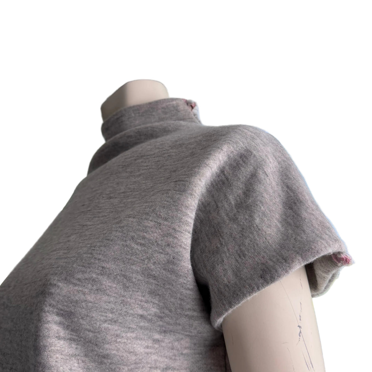 A close-up of a light gray knit turtleneck sweater draped on a mannequin torso, positioned in front of a window with a view of blue sky and greenery outside.